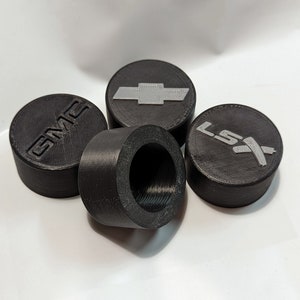 May include: Four black, cylindrical automotive parts with embossed logos. One features the GMC logo, another the Chevrolet bowtie, and a third displays the LSX emblem. The fourth part is hollow, revealing a threaded interior.