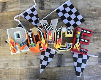Puede incluir: Decoraciones coloridas con temática de carreras que deletrean la palabra "RACE". Las letras están decoradas con imágenes de coches de carreras, llamas y banderas a cuadros. Incluye un semáforo y un trofeo.