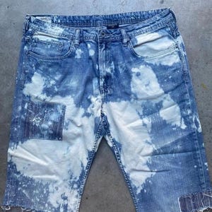 Puede incluir: Pantalones cortos de mezclilla desgastados con un diseño salpicado de lejía. Los pantalones cortos tienen un dobladillo deshilachado y un parche en la pierna izquierda.