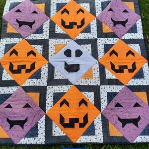 Op de afbeelding: Een Halloween-quilt met herhalende ruitvormen met pompoen- en spookgezichten. De quilt heeft een zwarte rand en ligt op een groene grasachtergrond. De quilt heeft oranje, paarse, grijze en witte kleuren.