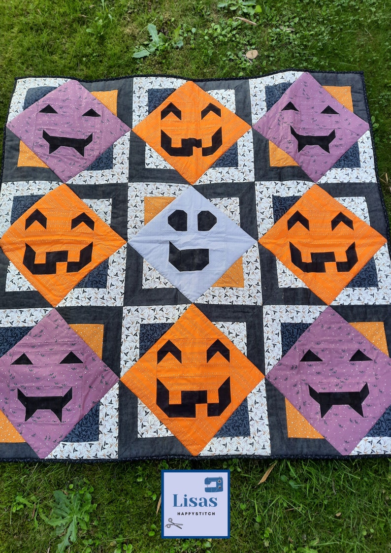 Op de afbeelding: Een Halloween-quilt met herhalende ruitvormen met pompoen- en spookgezichten. De quilt heeft een zwarte rand en ligt op een groene grasachtergrond. De quilt heeft oranje, paarse, grijze en witte kleuren.