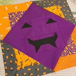 Op de afbeelding: Een Halloween-quiltblok met een paars pompoengezicht, zwarte driehoekige ogen en een grillige mond. Het blok is omgeven door oranje en grijze stof met Halloween-patronen, waaronder geesten, vleermuizen en pompoenen. De quilt ligt op een snijmat.
