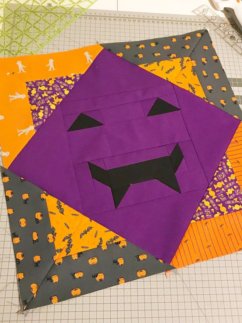 Op de afbeelding: Een Halloween-quiltblok met een paars pompoengezicht, zwarte driehoekige ogen en een grillige mond. Het blok is omgeven door oranje en grijze stof met Halloween-patronen, waaronder geesten, vleermuizen en pompoenen. De quilt ligt op een snijmat.