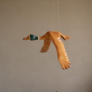 Puede incluir: M&oacute;vil de madera con forma de pato en vuelo, suspendido por finos hilos. El pato tiene un acabado de madera natural con cabeza verde y detalles blancos en las alas. Las alas est&aacute;n extendidas, dando la impresi&oacute;n de movimiento.