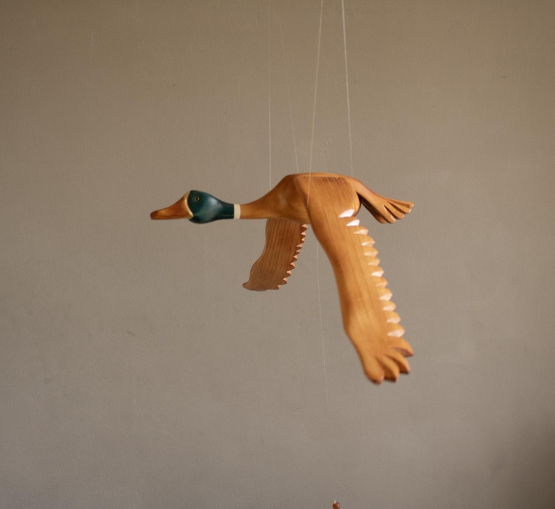 Puede incluir: M&oacute;vil de madera con forma de pato en vuelo, suspendido por finos hilos. El pato tiene un acabado de madera natural con cabeza verde y detalles blancos en las alas. Las alas est&aacute;n extendidas, dando la impresi&oacute;n de movimiento.