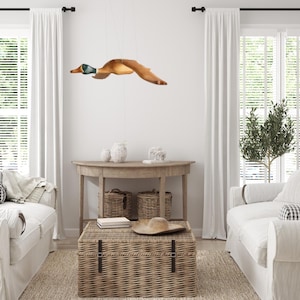 Puede incluir: Una l&aacute;mpara con forma de pato de madera cuelga del techo. El pato es marr&oacute;n con la cabeza verde y azul, con las alas extendidas en vuelo. La habitaci&oacute;n cuenta con sof&aacute;s blancos, un ba&uacute;l tejido y una mesa de madera.