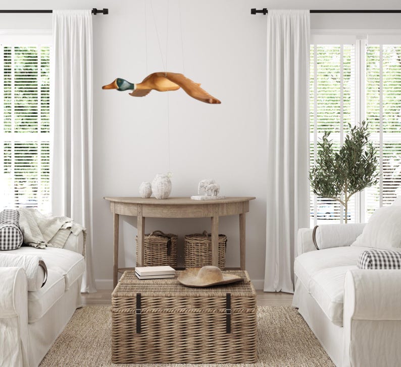 Puede incluir: Una l&aacute;mpara con forma de pato de madera cuelga del techo. El pato es marr&oacute;n con la cabeza verde y azul, con las alas extendidas en vuelo. La habitaci&oacute;n cuenta con sof&aacute;s blancos, un ba&uacute;l tejido y una mesa de madera.