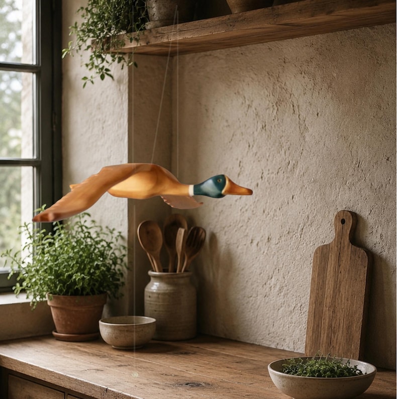 Puede incluir: Un m&oacute;vil de madera con forma de pato, pintado en tonos marr&oacute;n, blanco y azul, cuelga en una cocina. El pato est&aacute; suspendido por un hilo casi invisible. La cocina cuenta con una encimera de madera, una tabla de cortar y plantas en macetas.