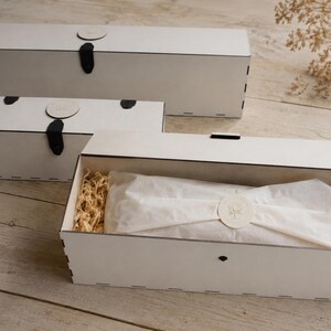 Puede incluir: Tres cajas de madera blancas con cierres negros y etiquetas redondas blancas. Una caja est&aacute; abierta, revelando un art&iacute;culo envuelto y material de embalaje. Las cajas est&aacute;n sobre una superficie de madera clara.