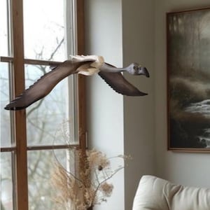 May include: A wooden bird sculpture in flight, with outstretched wings. The bird has a gray head and a dark brown body and wings. The sculpture is positioned in front of a window and a painting.