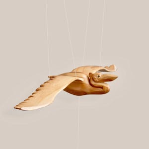 Könnte beinhalten: Eine Holzskulptur eines Vogels im Flug, aufgehängt an dünnen weißen Schnüren. Der Vogel ist aus hellem Holz geschnitzt, mit detaillierten Flügeln und einem langen Schnabel. Die Skulptur befindet sich vor einem neutralen Hintergrund.