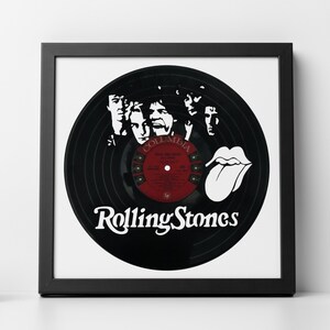 May include: A black vinyl record with a white silhouette of the Rolling Stones band members and the band's name in white lettering. The record has a red and white Columbia Records label with the album title "Sticky Fingers" printed on it. The record is framed in a black frame.