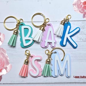Könnte beinhalten: Schlüsselanhänger mit den Buchstaben B, A, K, S und M in verschiedenen Farben, jeweils mit einem passenden Quastenanhänger. Die Buchstaben sind weiß mit farbigem Rand und gewelltem Rand. Goldfarbene Schlüsselringe und Quastenabschlüsse runden das Design ab.