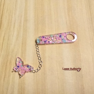 May include: A pink and white butterfly charm with a silver chain attached to a pink and white glitter rectangle with a hole in the top. The charm is on a wooden surface.  Laser Authority.
