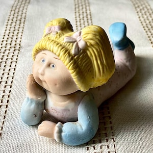 May include: A porcelain figurine of a baby lying down, with blonde hair and blue eyes. The baby is wearing a light pink outfit with blue accents on the sleeves and feet. The figurine is resting on a cream-colored surface.