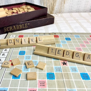 May include: A close-up of a Scrabble game board with tiles spelling out the words "D,N,I,V,L" and "T,E,D,G,A,P". The tiles are on wooden racks and the board is made of a light brown material.