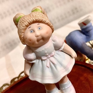 May include: A porcelain figurine of a little girl with blonde hair, wearing a light blue dress with a pink bow and white shoes. She has rosy cheeks and big blue eyes.