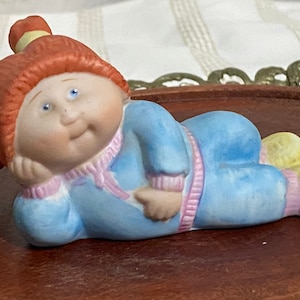 May include: A ceramic figurine of a child in a light blue onesie with pink trim, lying on a wooden surface. The child has red hair and yellow accents. The figurine is a collectible item.