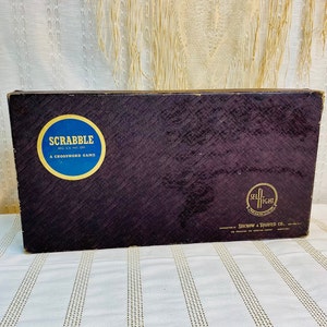 May include: A vintage Scrabble game box with a dark brown textured surface. The box features a blue and gold circular logo with the word "SCRABBLE" and the text "REG. U.S. PAT. OFF. A CROSSWORD GAME". The bottom of the box has a gold circular logo with the text "SELRIGHT" and "SELCHOW & RIGHTER CO."