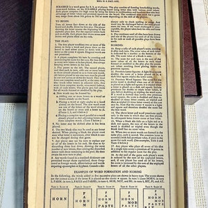 May include: Close-up of the back of a Scrabble game box with the rules for playing printed on a cream-colored background. The rules include instructions for scoring, forming words, and using premium squares. The box is made of brown cardboard with a cream-colored interior.