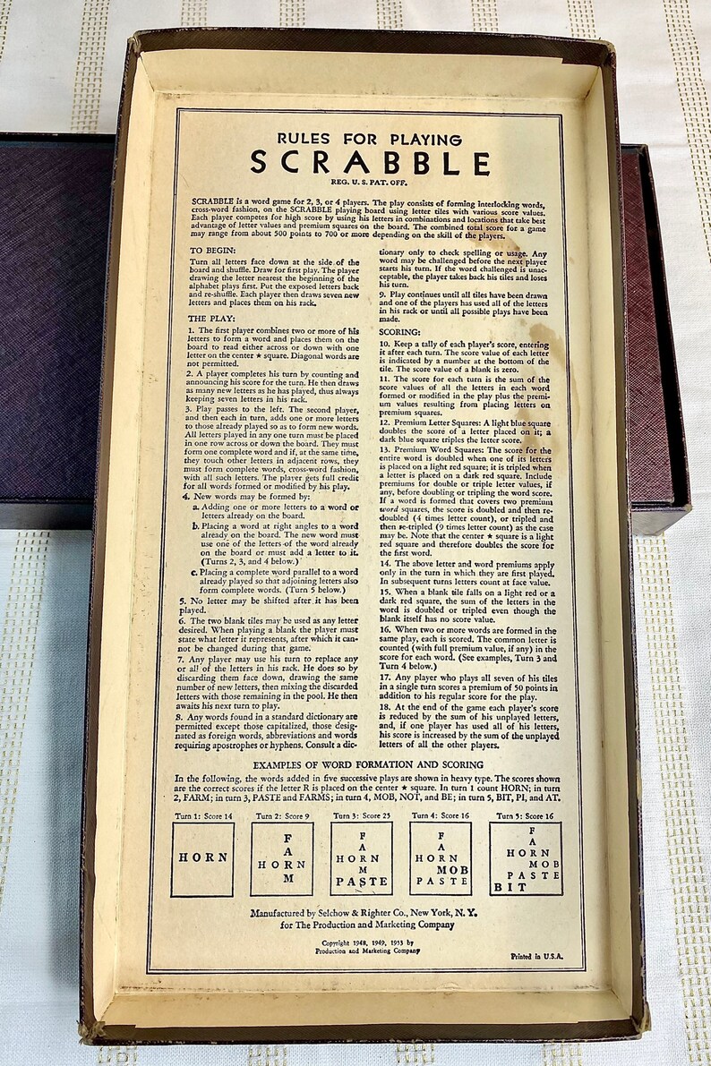 May include: Close-up of the back of a Scrabble game box with the rules for playing printed on a cream-colored background. The rules include instructions for scoring, forming words, and using premium squares. The box is made of brown cardboard with a cream-colored interior.