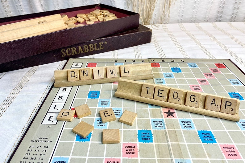 May include: A close-up of a Scrabble game board with tiles spelling out the words "D,N,I,V,L" and "T,E,D,G,A,P". The tiles are on wooden racks and the board is made of a light brown material.