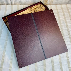 May include: A burgundy-colored wooden box with a hinged lid. The box is open and filled with wooden letter tiles. The tiles are arranged in a random order. The box is made of a textured wood.