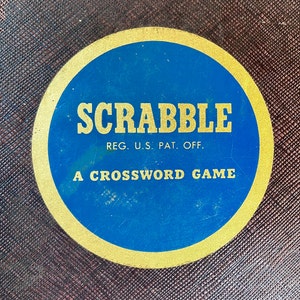 May include: A close-up of a gold and blue circular game board with the text "SCRABBLE REG. U.S. PAT. OFF. A CROSSWORD GAME" printed on it.