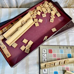 May include: A wooden box filled with letter tiles for a Scrabble game. The tiles are scattered inside the box, with some letters visible, such as 'A', 'E', 'S', and 'J'. The box is open and the lid is lying on a white surface with a striped pattern.