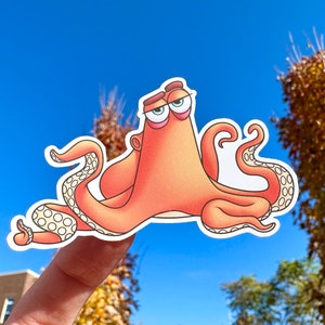 May include: A sticker of an orange octopus with large eyes and a sad expression. The octopus has seven tentacles with white spots. The sticker is on a finger against a blue sky background.