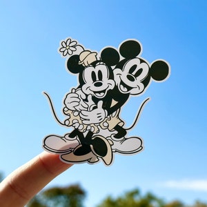 May include: Black and white sticker of Mickey Mouse and Minnie Mouse hugging, with Minnie Mouse wearing a flower on her head. The sticker is on a finger against a blue sky background.