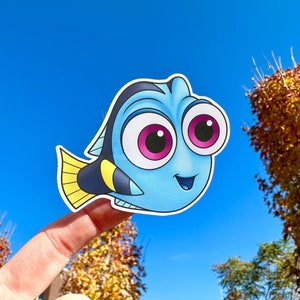 May include: A blue and yellow cartoon fish with big eyes and a smile. The fish is holding a finger up in the air, as if it is pointing at something.
