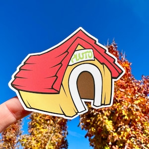 May include: A yellow and red cartoon dog house with the word "Pluto" written on the roof. The dog house is being held up by a finger against a blue sky and fall foliage background.