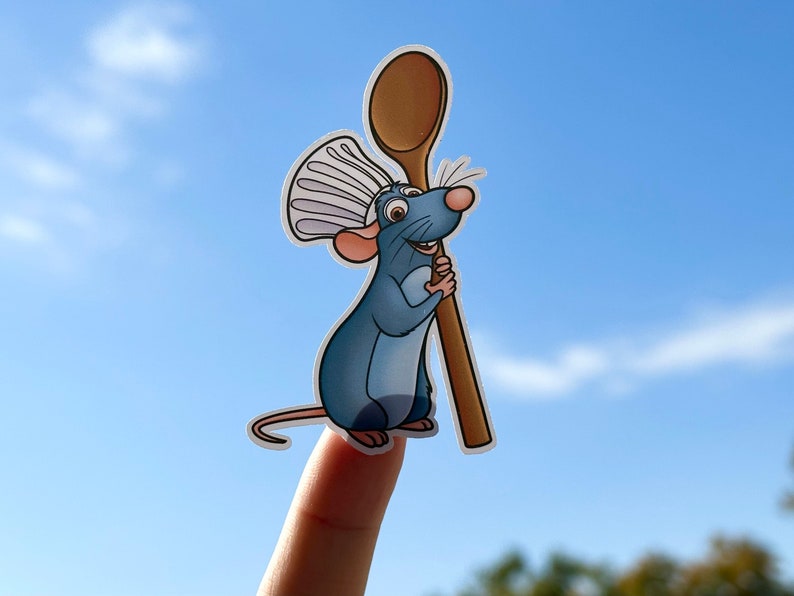 K&ouml;nnte beinhalten: Eine blaue Comic-Ratte mit Kochm&uuml;tze h&auml;lt einen Holzl&ouml;ffel. Die Ratte l&auml;chelt und hat einen fr&ouml;hlichen Gesichtsausdruck. Der Hintergrund ist ein blauer Himmel mit wei&szlig;en Wolken.