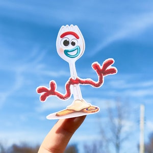 May include: A white plastic fork with red and blue details, shaped like a cartoon character with a smiling face. The character has red pipe cleaner arms and is standing on a finger.