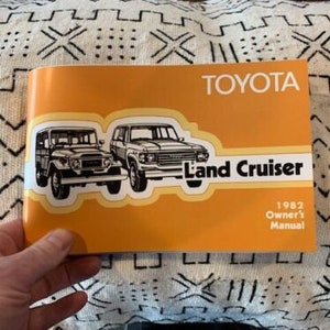 May include: An orange Toyota Land Cruiser owner's manual from 1982. The cover features illustrations of two Land Cruiser models in black and white. The Toyota logo is at the top, with "Land Cruiser" in a white banner.