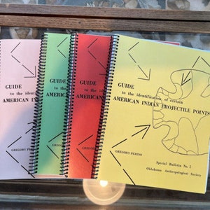May include: Four spiral bound guide books with yellow, pink, green, and red covers. The covers have black text and arrows. The yellow book has the title "GUIDE to the identification of certain AMERICAN INDIAN PROJECTILE POINTS" and "Special Bulletin No. 2 Oklahoma Anthropological Society".