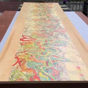 Puede incluir: Una mesa de madera rectangular grande con un diseño abstracto colorido. La obra de arte presenta líneas arremolinadas en tonos de rojo, amarillo, verde y azul. Los bordes de la mesa son de color marrón claro y el diseño cubre la mayor parte de la superficie.