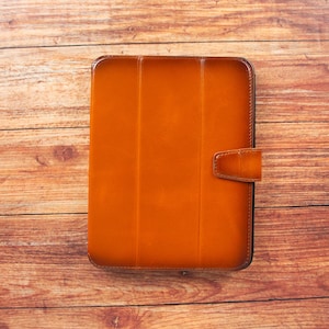 May include: A rectangular, orange-brown leather tablet case with a magnetic closure. The case is on a wooden surface with a natural grain pattern. The case has a vertical design with three panels.
