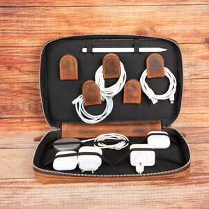 May include: Open leather tech organizer with a black interior, holding white charging cables, a stylus, and white charging blocks. The organizer has brown leather cable holders and a zipper closure. The case is open, revealing its contents.