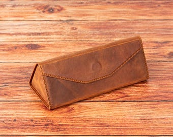 May include: A triangular-shaped brown leather glasses case with a pointed flap closure. The case features visible tonal stitching along the edges and a smooth, matte finish, designed to protect eyewear with its structured, rigid form.