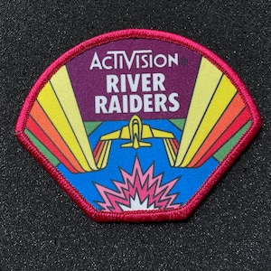 May include: A red, white, and blue patch with a rainbow design and a plane flying over a pink explosion. The patch says "Activision River Raiders" in white text.