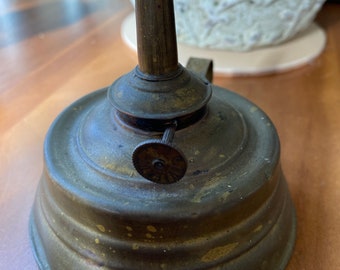 Puede incluir: Una lámpara de aceite antigua de latón con una base redonda y un portamechas cilíndrico alto. La lámpara tiene un pequeño pomo redondo en la parte superior para ajustar la mecha.