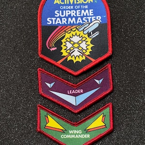 May include: Set of three embroidered patches. The top patch features the Activision logo and the text "ORDER OF THE SUPREME STARMASTER". Below are two chevron-shaped patches with the text "LEADER" and "WING COMMANDER". All patches have red borders.