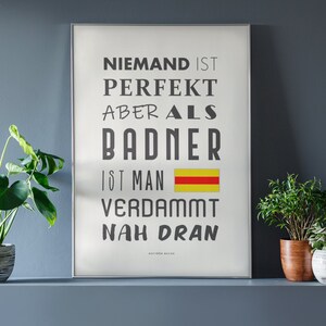 May include: A white framed poster with black text that reads "Niemand ist perfekt aber als Badner ist man verdammt nah dran". The poster features a yellow and red stripe in the middle. The poster is hanging on a grey wall with three potted plants in front of it.
