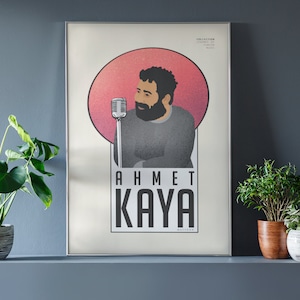 May include: A framed poster with a minimalist design featuring a man with a beard and a microphone in front of a pink circle. The text "AHMET KAYA" is printed below the image.