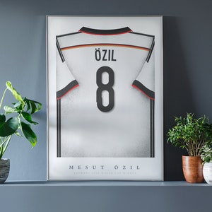 May include: A framed poster of a white soccer jersey with black stripes and the name "Özil" and number "8" on the back. The poster also includes the text "Mesut Özil" and "German FIFA World Cup Winner".