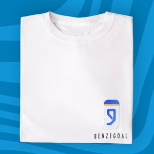 Może przedstawiać: Biała koszulka z haftowanym logo w kolorze niebieskim i ż&oacute;łtym, na kt&oacute;rym widnieje liczba "9" i napis "BENZEGOAL" poniżej.