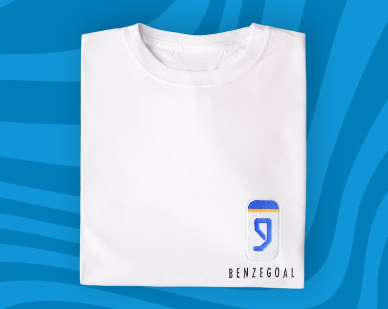 Może przedstawiać: Biała koszulka z haftowanym logo w kolorze niebieskim i ż&oacute;łtym, na kt&oacute;rym widnieje liczba "9" i napis "BENZEGOAL" poniżej.