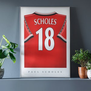 May include: A framed poster featuring a red soccer jersey with black and white trim, displaying the name SCHOLES and the number 18 in white. Below the jersey, the text reads PAUL SCHOLES and MANCHESTER UNITED CHAMPIONS LEAGUE WINNER 98-99.